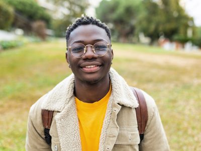Smiling african young student man looking at camera outdoors - Cheerful 20s guy with backpack standing at university campus